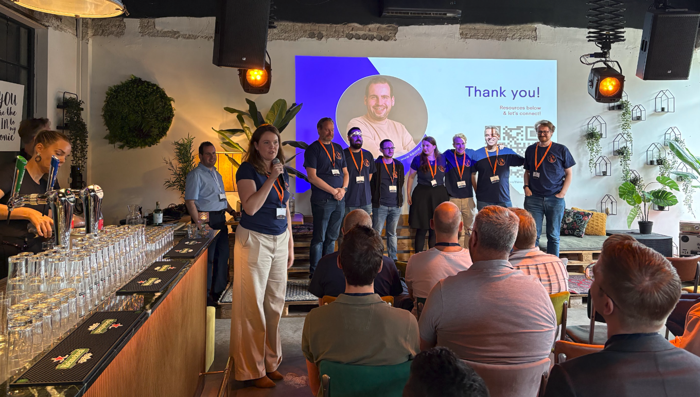 A speaker addresses an audience while a smiling event team stands before a “Thank you!” screen in a lively, warmly lit venue.