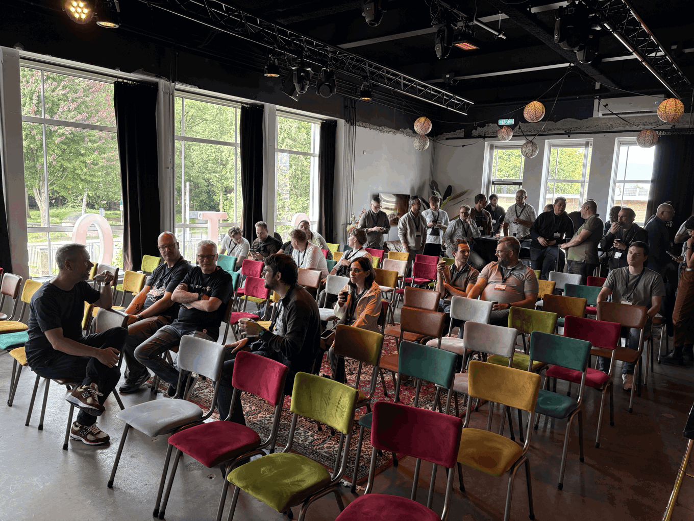 A diverse audience gathers in a bright event room with colorful chairs, large windows, and hanging lights, creating a relaxed, welcoming atmosphere.